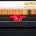 A close-up shot of a bright red plastic mounting bracket or handle attached to a black metal surface, with a blurred background showing warm orange and white tones.